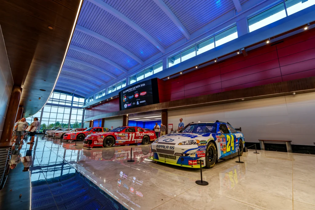 Row of NASCAR race cars indoors with reflective tiled floor and large windows showing outside trees