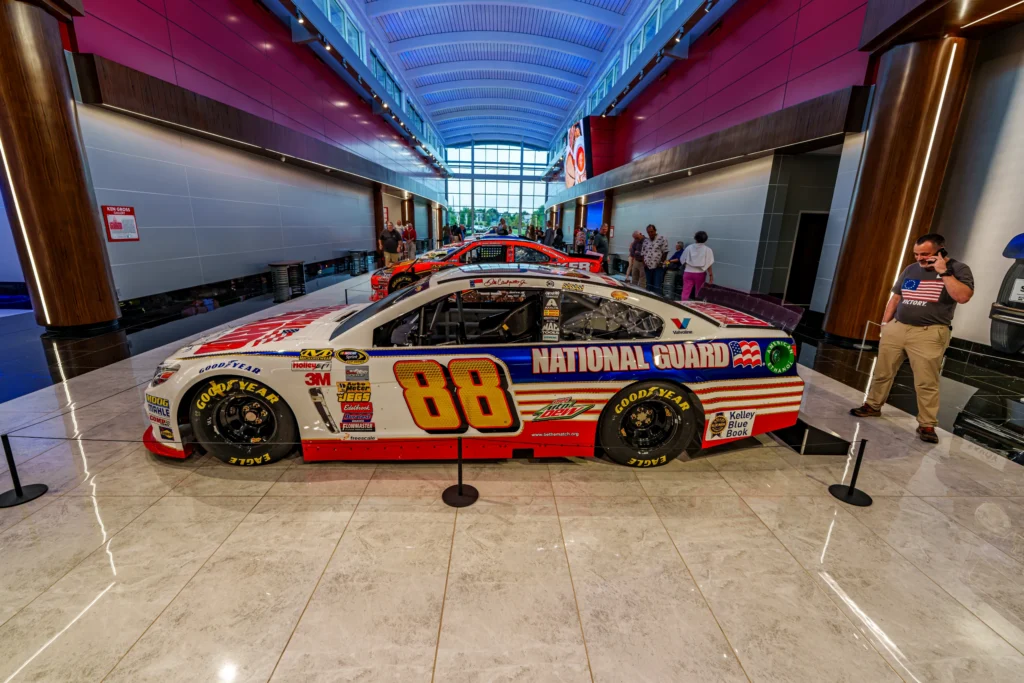 White and red NASCAR stock cars displayed indoors with people standing nearby in a gallery setting.