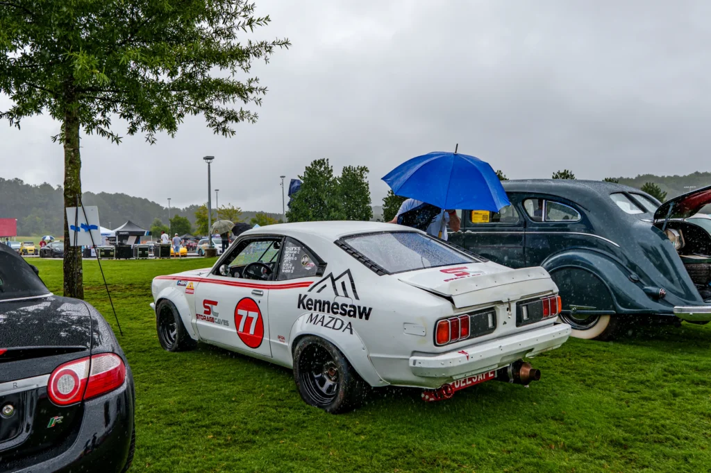 White Mazda RX-3 with racing decals parked on grass at a rainy outdoor car event with other vintage cars and umbrellas