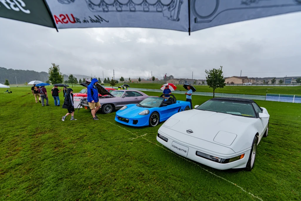 White Chevrolet Corvette convertible, blue Lotus Elise convertible, and gray classic coupe parked on wet grass with people