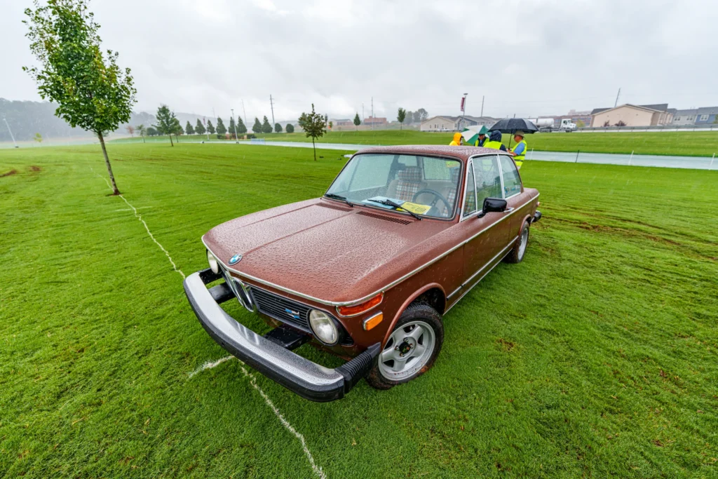 Brown BMW 2002 parked on wet grass with rain falling and a group of people with umbrellas in the background
