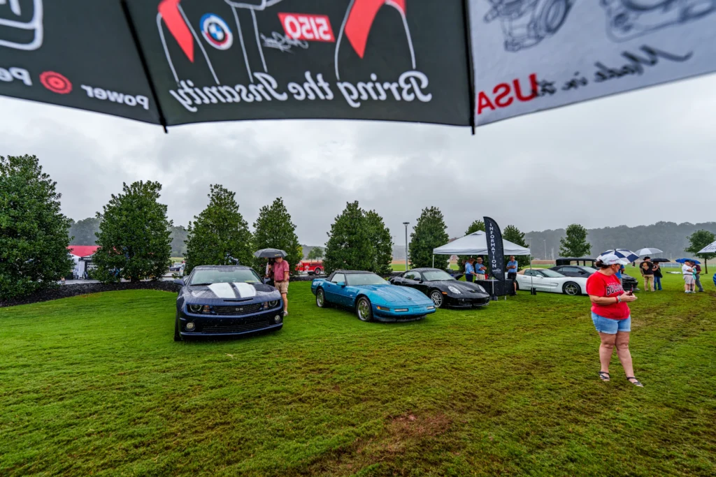 Three cars on grass in rainy weather with people holding umbrellas and a tent and trees in the background.