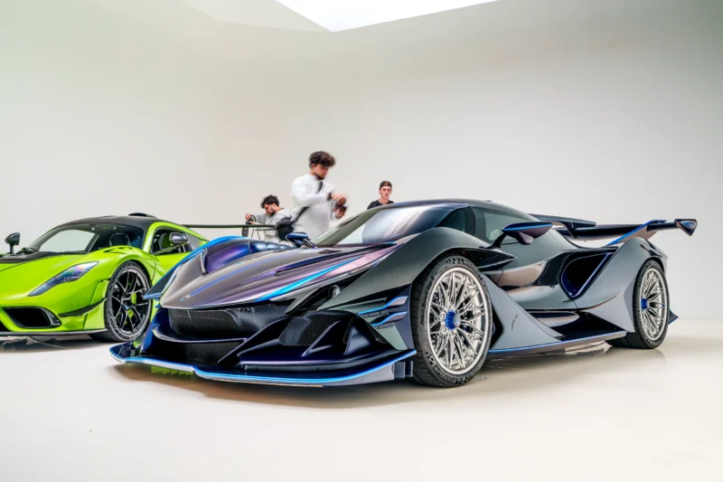 Two parked sports cars indoors with people walking behind them; foreground car is black with blue accents, background car is