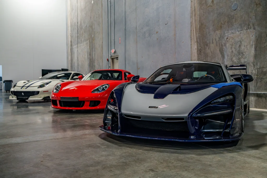 Three high-performance sports cars parked indoors against a concrete wall background, white Ferrari, red Porsche, and silver
