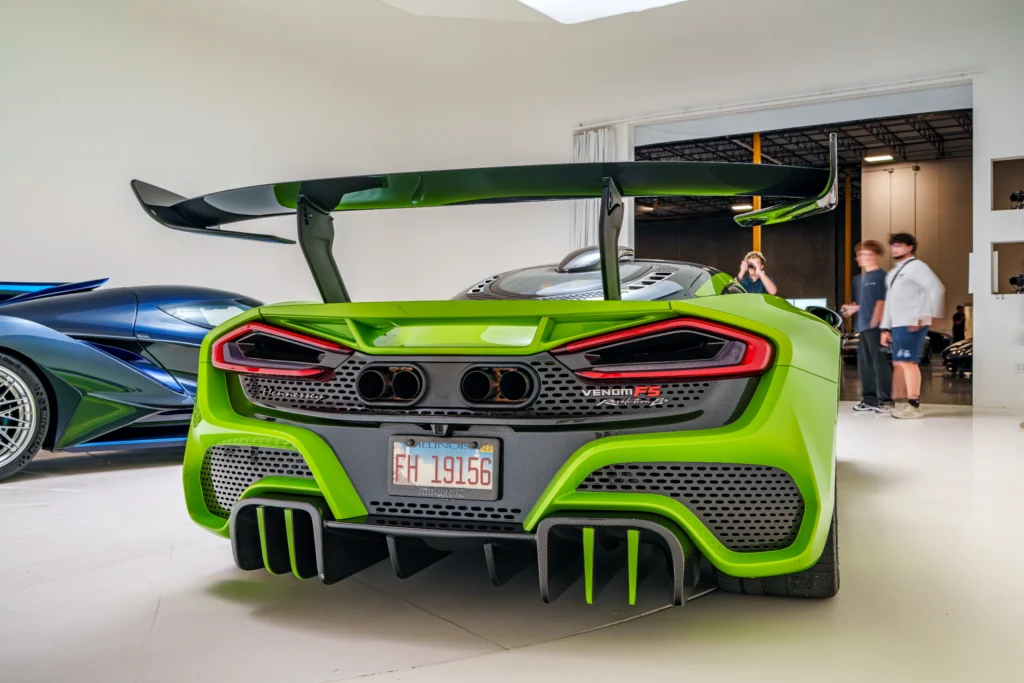 Rear view of a green Hennessey Venom F5 Revolution Evo with a large spoiler inside a showroom with a blue sports car nearby