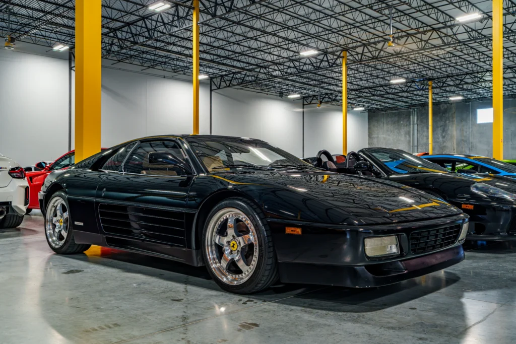 Black Ferrari 348 TS parked indoors with other sports cars in the background under a high ceiling with yellow support beams