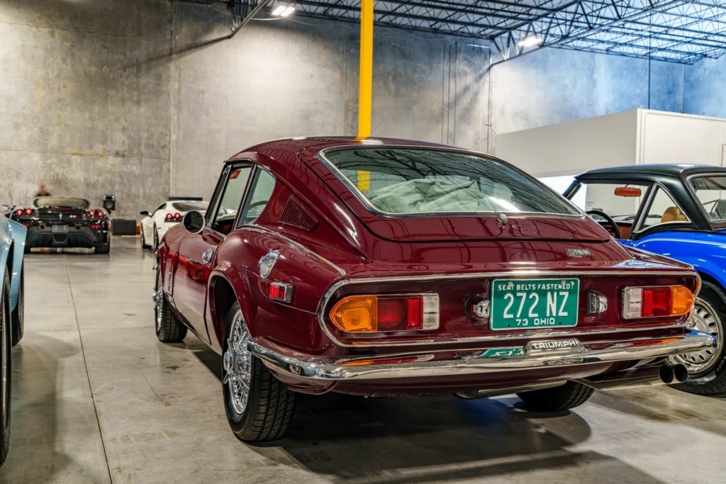 Burgundy Triumph GT6 car rear view in an indoor garage with other vehicles nearby under industrial lighting