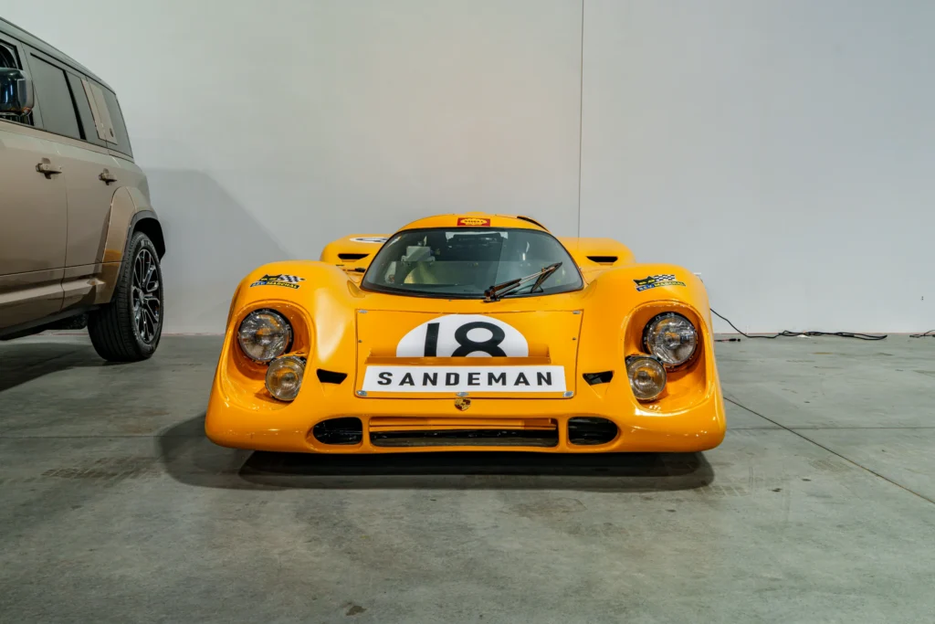 Yellow Porsche race car with number 18 and Sandeman logo in indoor setting, partial SUV next to it