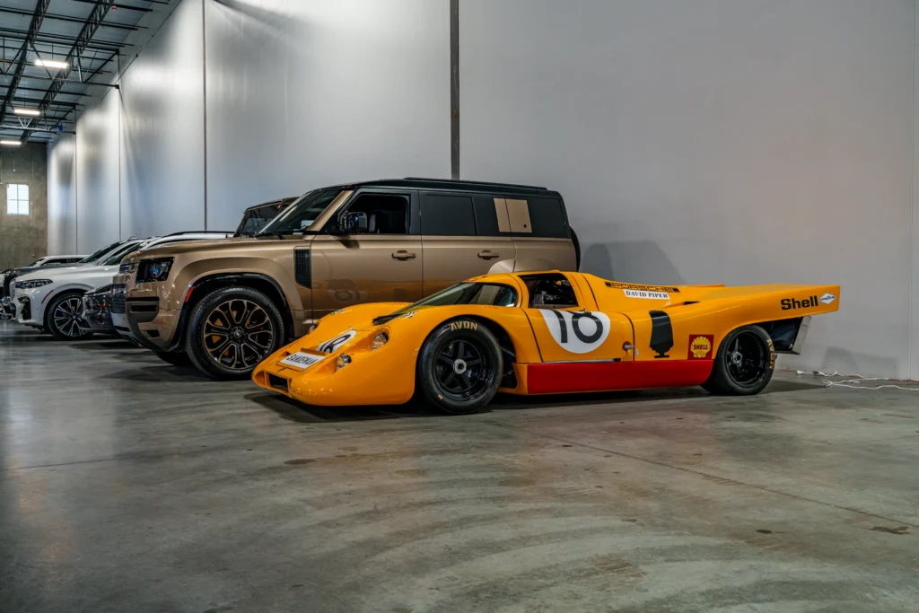 Yellow Porsche 917 race car parked inside with a brown Land Rover Defender and other cars in the background.