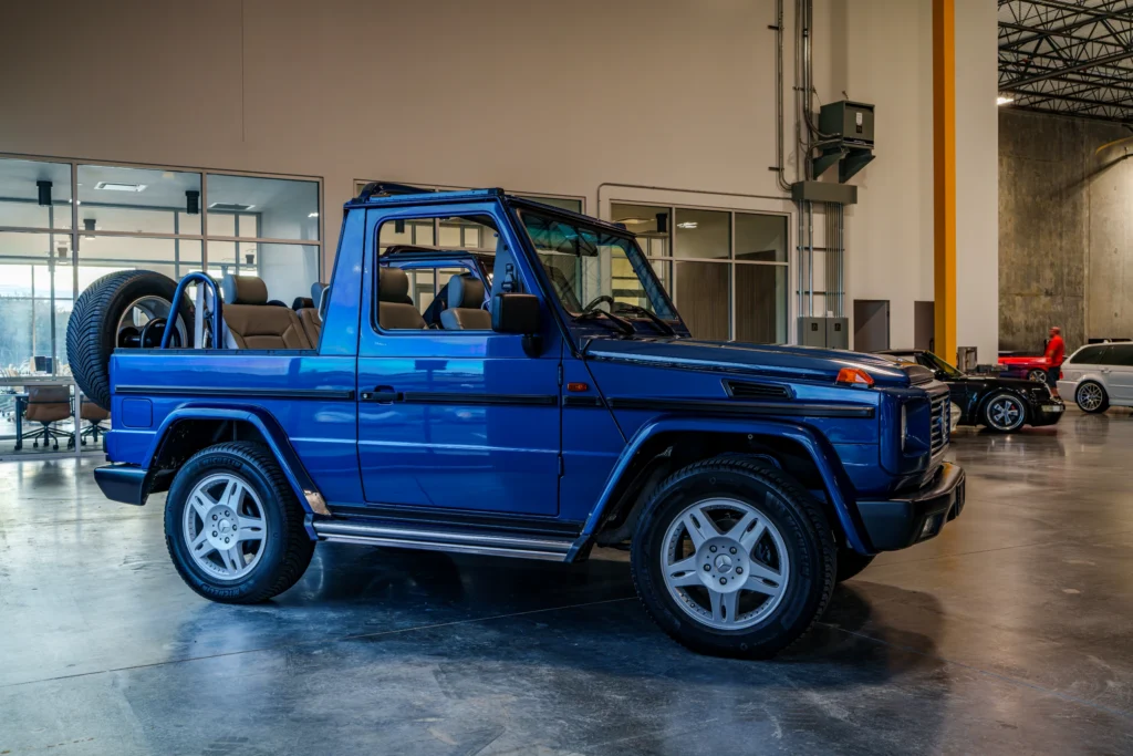 Blue Mercedes-Benz Cabriolet parked indoors in a showroom or garage with other cars and a person in the background.