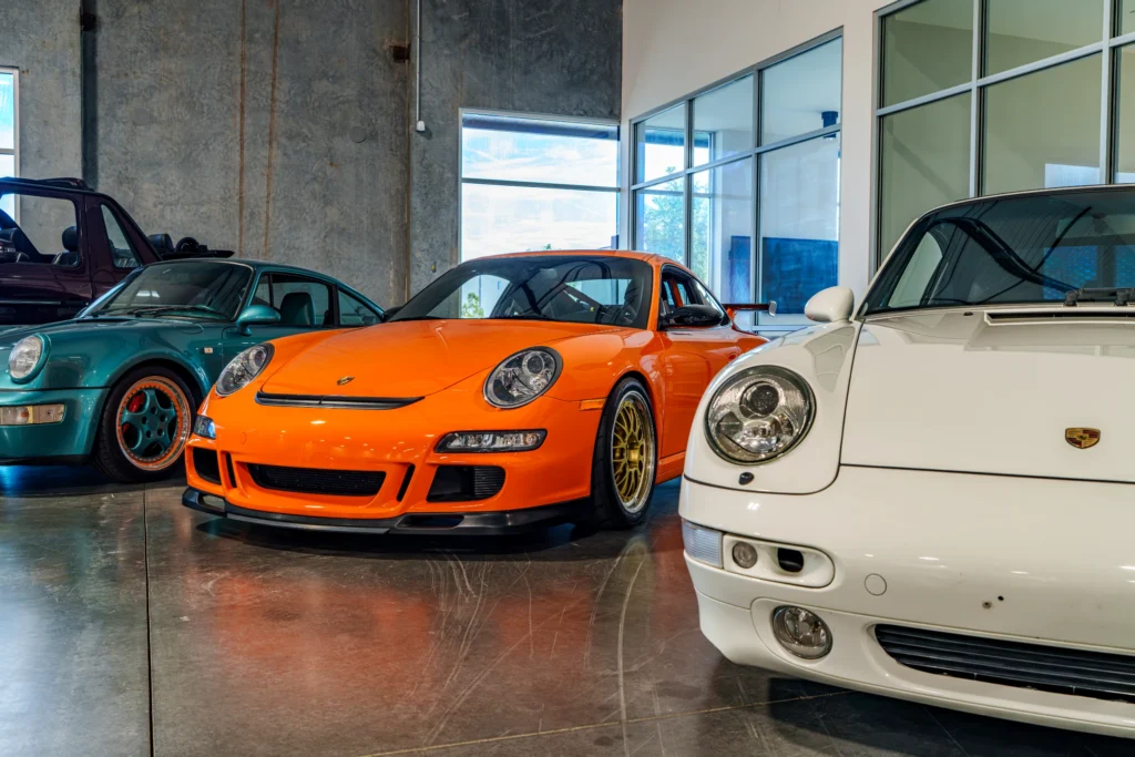 Three Porsche cars in a showroom, featuring a teal coupe, an orange coupe, and a white coupe, parked side by side indoors.