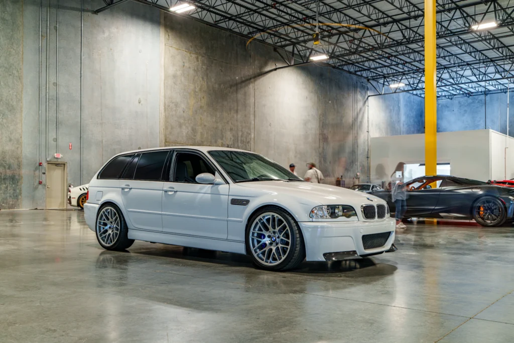 White BMW M3 wagon parked indoors with several other sports cars and people blurred in the background