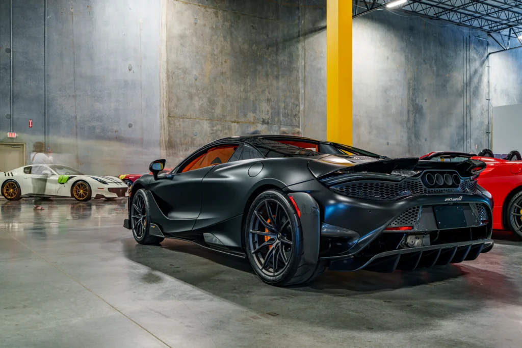 A black McLaren 765LT is displayed indoors, viewed from the rear three-quarter angle with other cars behind it.
