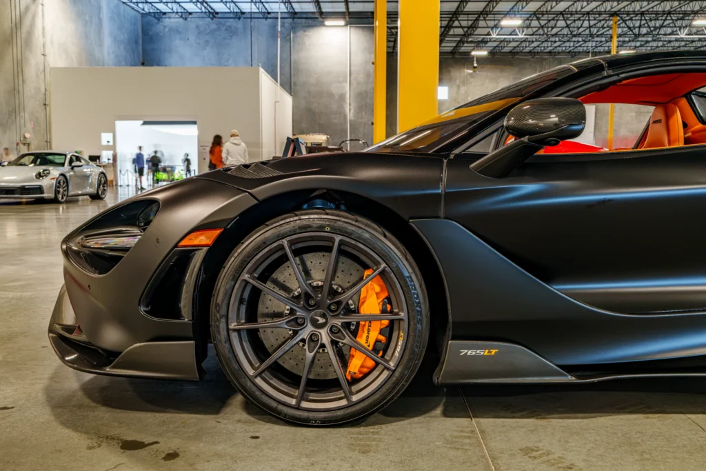 Black McLaren 765LT showing front wheel and side mirror inside a showroom with a silver Porsche 911 in the background