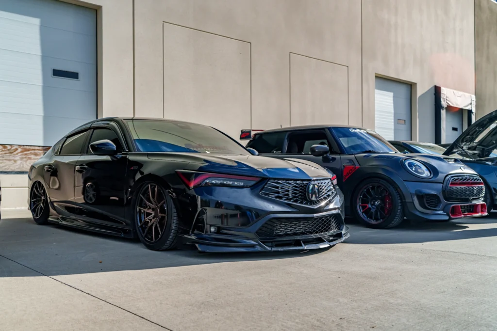 Black Acura Integra and blue Mini Cooper with decals parked side by side in front of a warehouse wall.