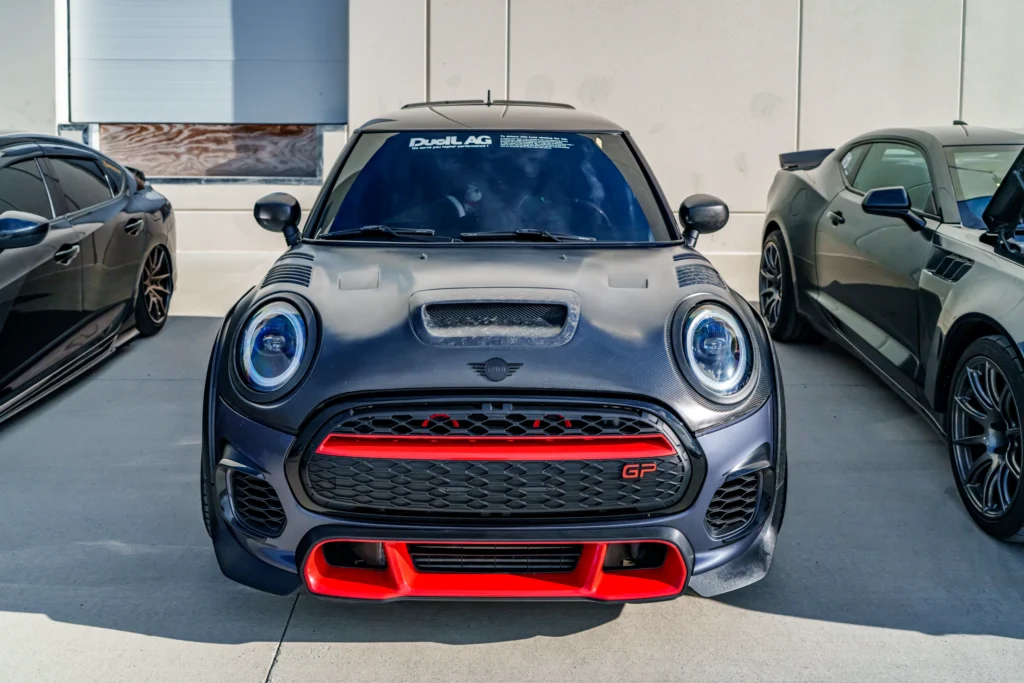 Front view of a black Mini Cooper GP parked between two other cars in an outdoor setting by a building wall.