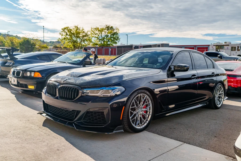 Black BMW sedan and dark blue BMW convertible parked outdoors at a car event with people and other vehicles in background.