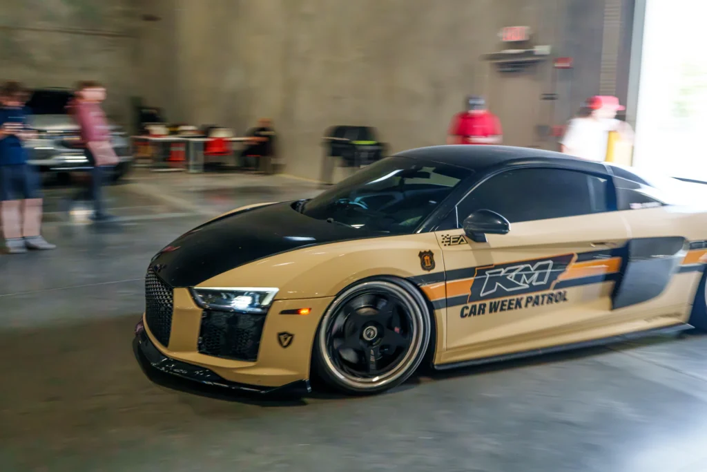 Tan and black Audi R8 with black wheels and decals parked indoors with blurred people in background