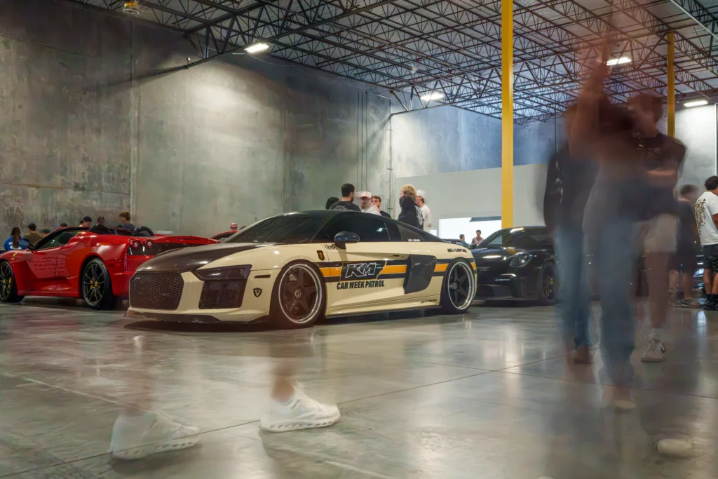 White and black Audi R8 with Car Week Patrol livery parked indoors with other sports cars and people walking nearby