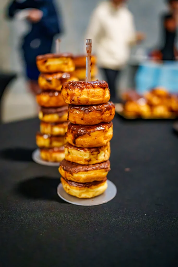 Stack of chocolate-glazed donuts on a stand with blurred people in the background.