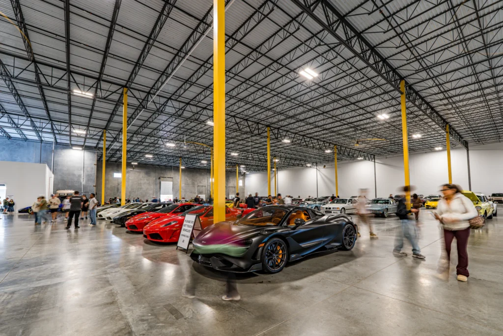 A black McLaren 765LT sits beside red Ferraris as attendees walk among rows of cars in a large exhibition hall.