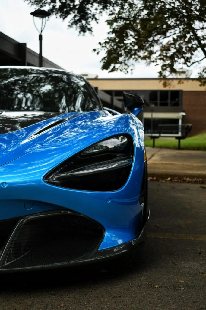 Front portion of blue McLaren 720S with distinct headlight design parked outdoors near a building and trees.