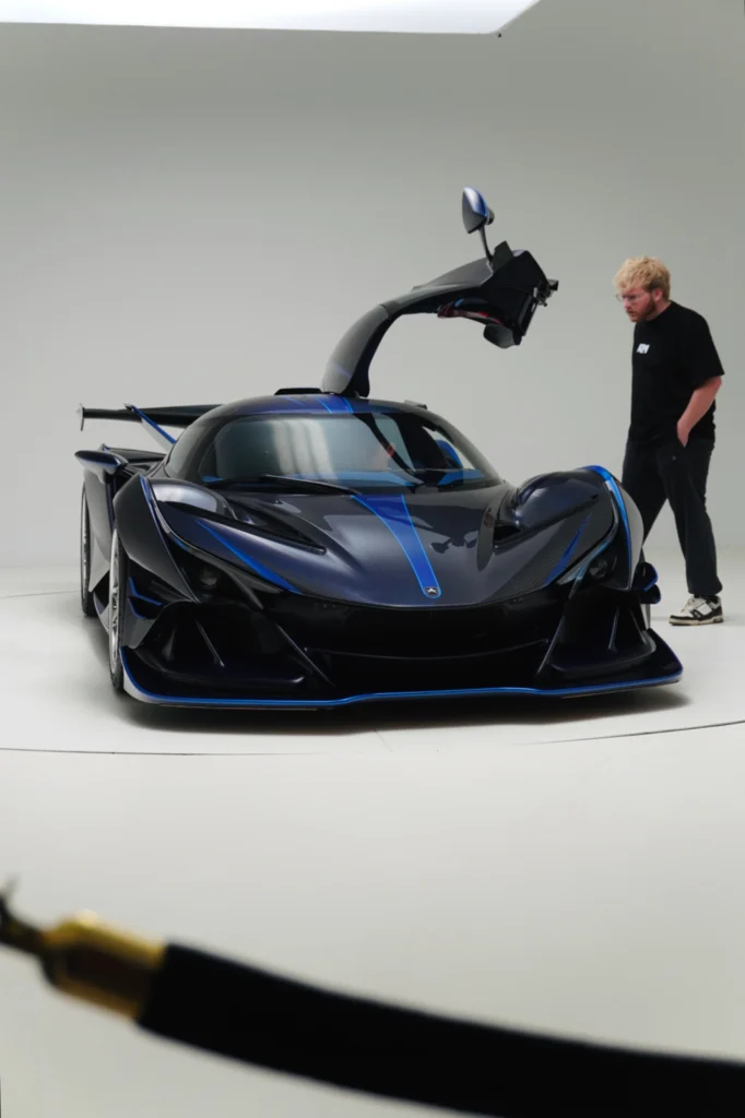 Black Apollo IE with blue accents and driver-side door open, indoor studio setting with person standing nearby