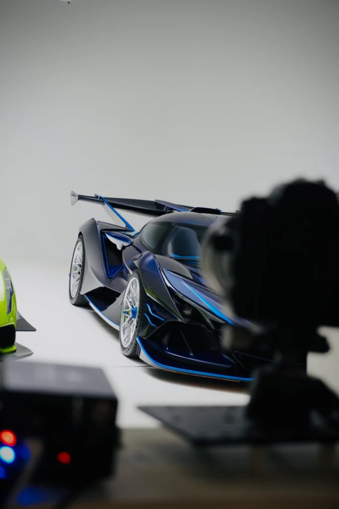 Black and blue Apollo IE with visible rear wing, parked indoors beside a green vehicle, with camera equipment in