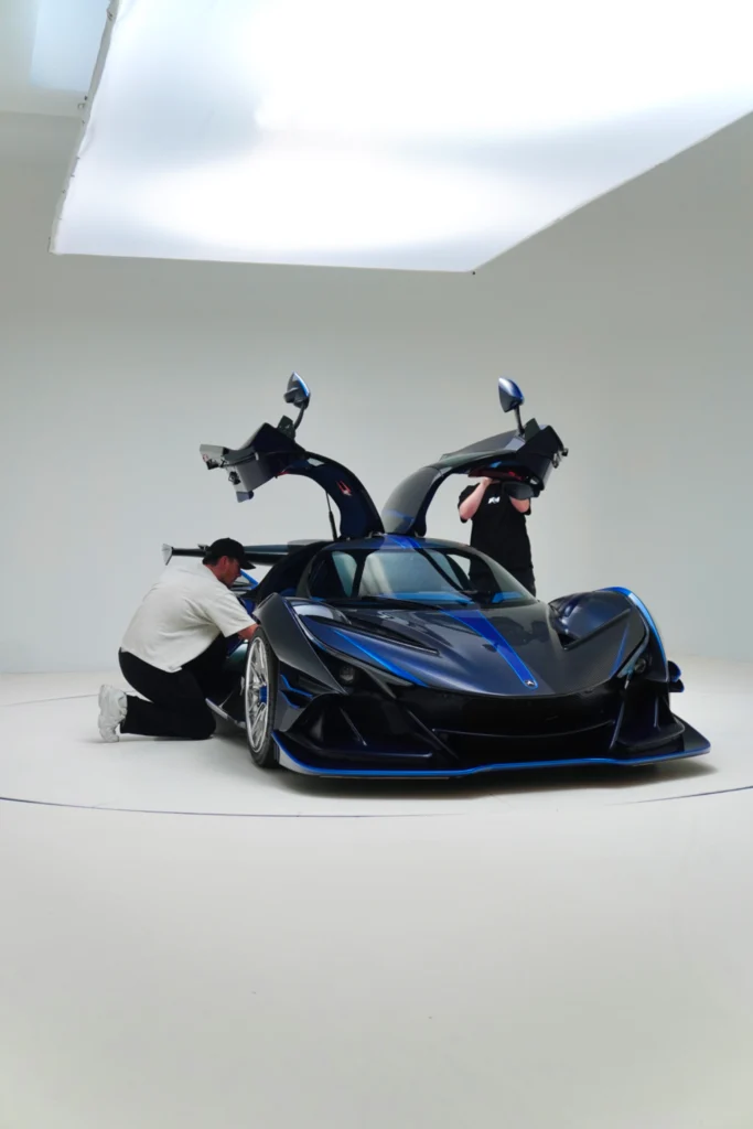 Blue Apollo IE with gullwing doors open and two men working on the car inside a white studio.