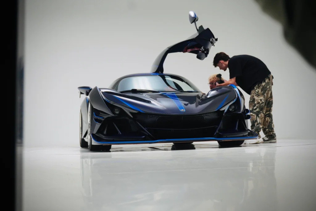 Blue and black Apollo IE with gullwing door open, two people examining the car, indoor studio setting