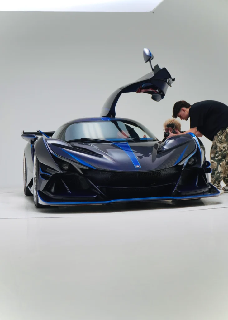 Blue and black Apollo IE with open driver-side butterfly door in a white studio with two people near the car.