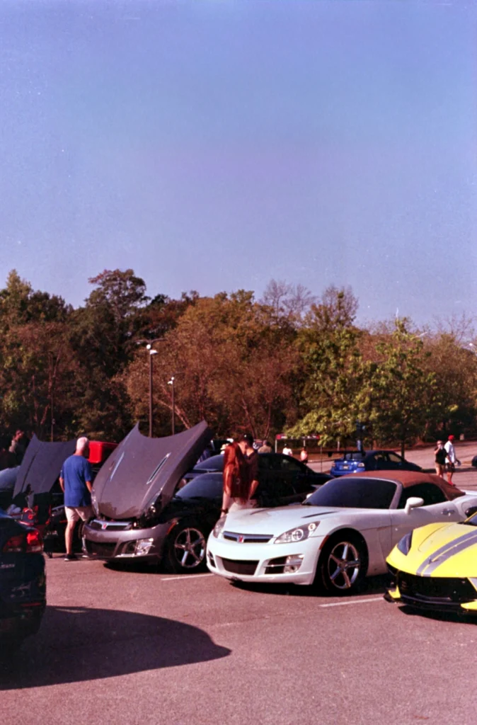 White Saturn Sky convertible with hood up and people nearby in a parking lot at an outdoor car event