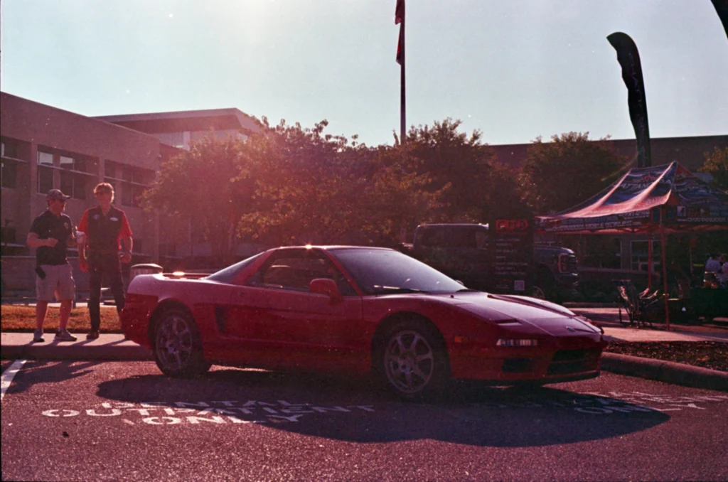 Red Acura NSX parked on asphalt with two men standing nearby and a building, trees, and event tent in background
