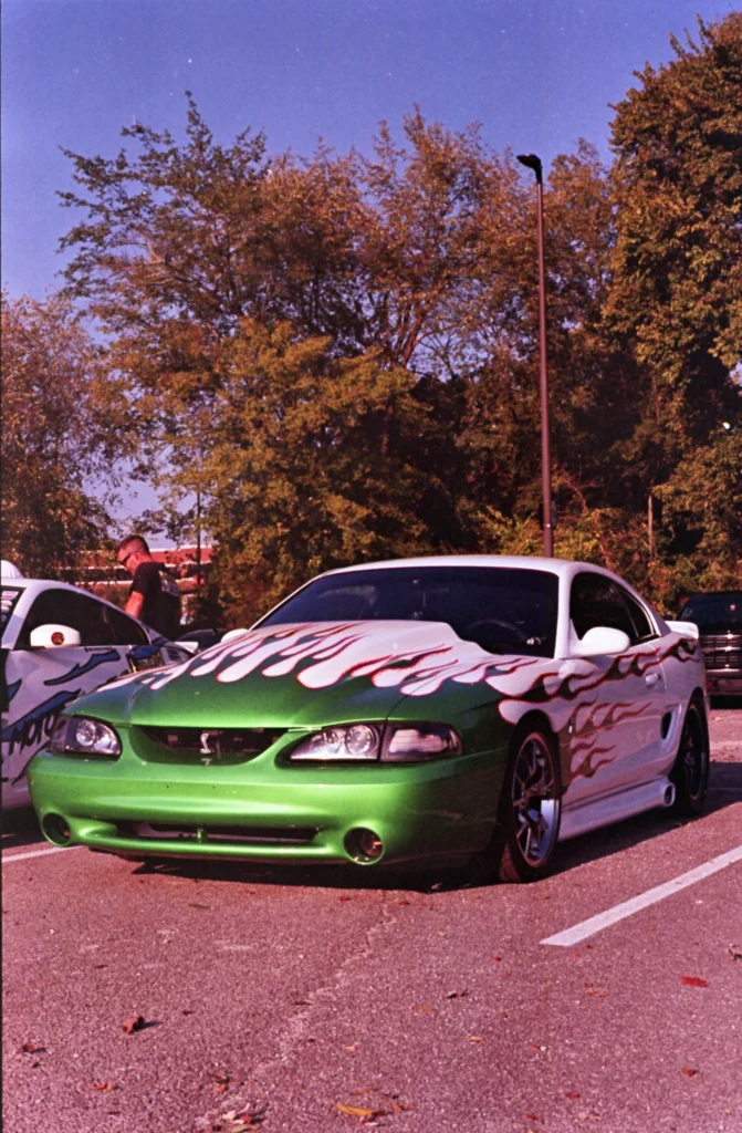 A white and green Ford Mustang with flame graphics is parked at an outdoor car show beneath trees.