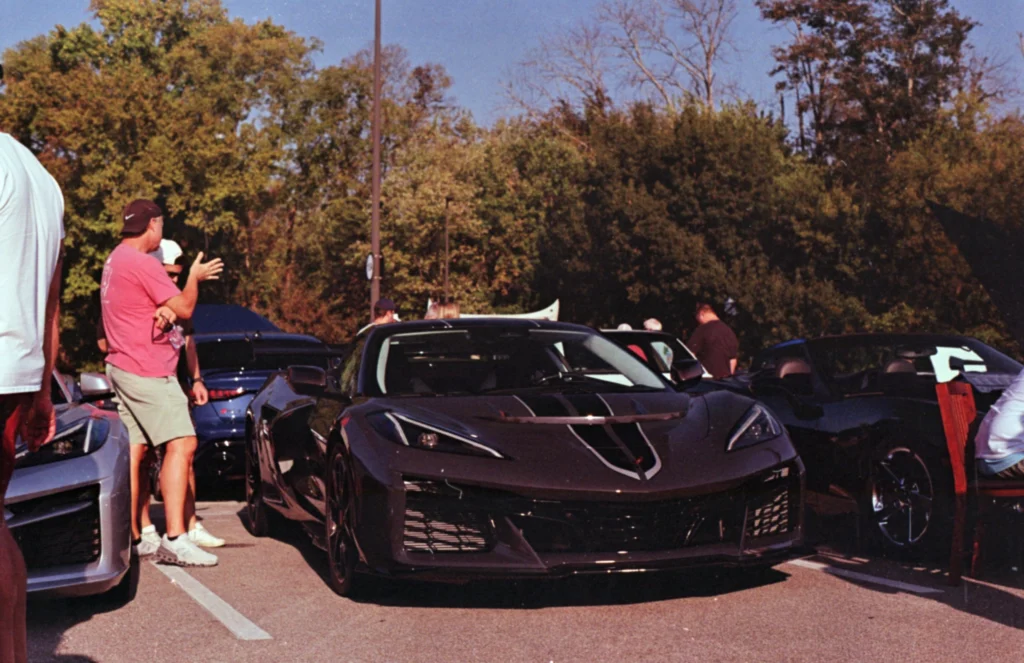 Black Chevrolet Corvette parked at a car event with people standing nearby and trees in the background