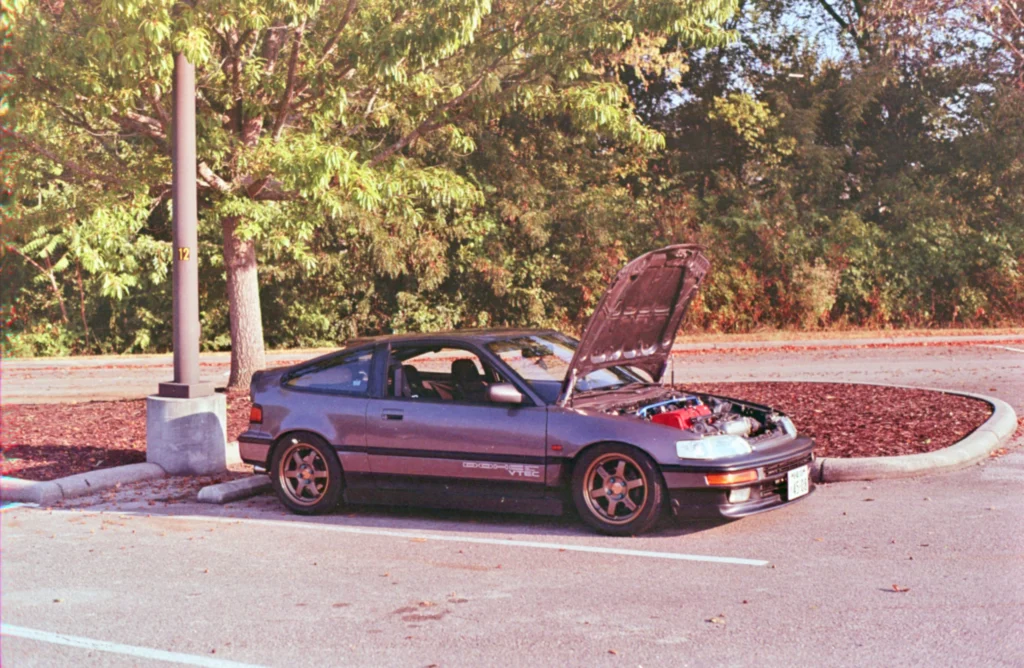 Gray Honda CRX with hood open parked in lot near trees and lamp post
