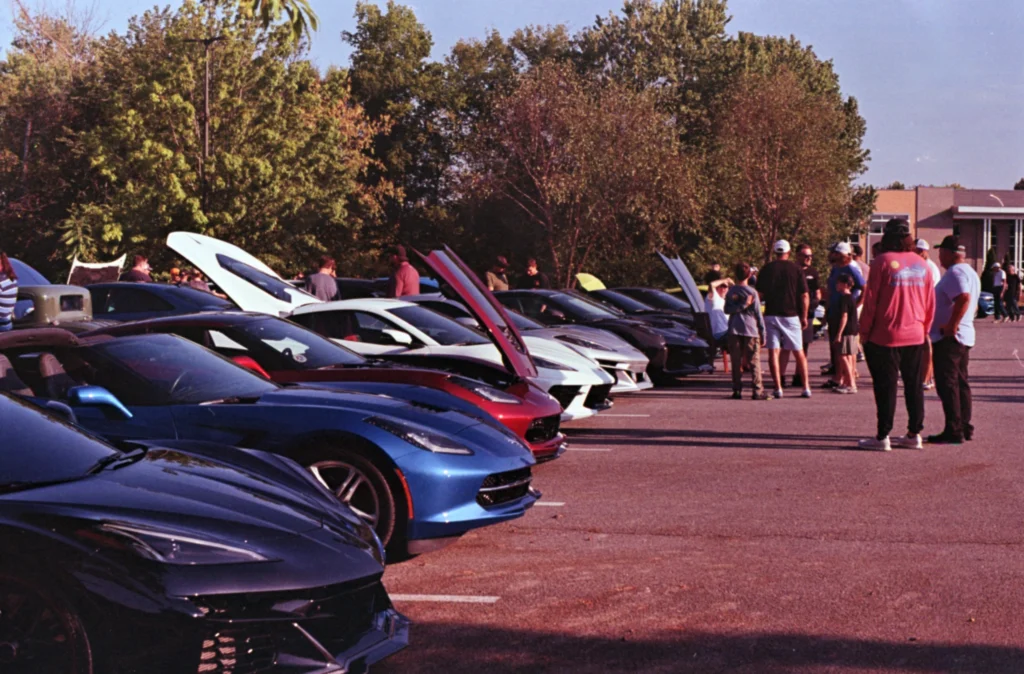 Row of Corvettes and Camaros with open hoods and attendees in a parking lot at a car event on a sunny day