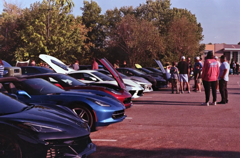 Row of Corvettes and Camaros with open hoods and attendees in a parking lot at a car event on a sunny day