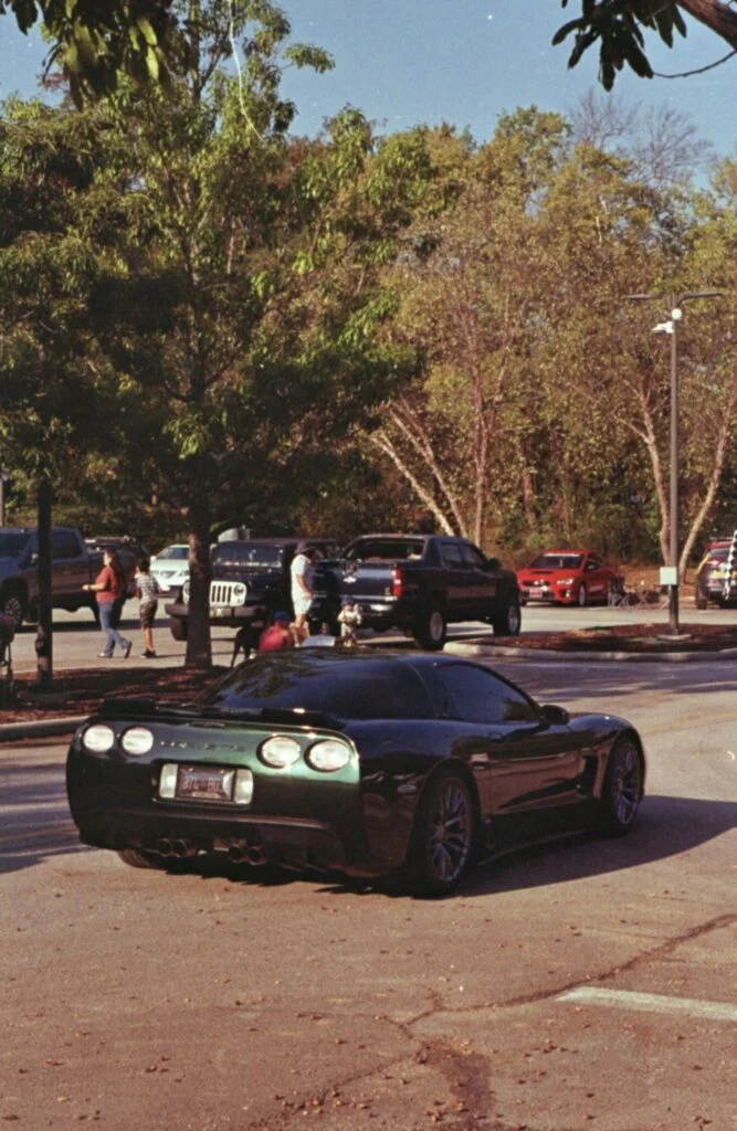 Dark green Chevrolet Corvette parked in a lot with trees and people in the background