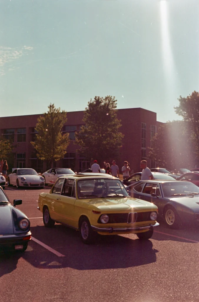 Yellow BMW 2002 parked with several other vehicles and people nearby in a lot with a building and trees in background