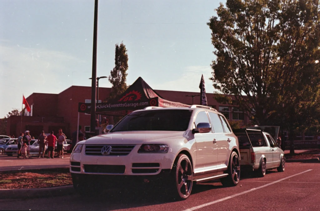A white Volkswagen Touareg sits ahead of a silver pickup at an outdoor car show with attendees and a vendor tent.