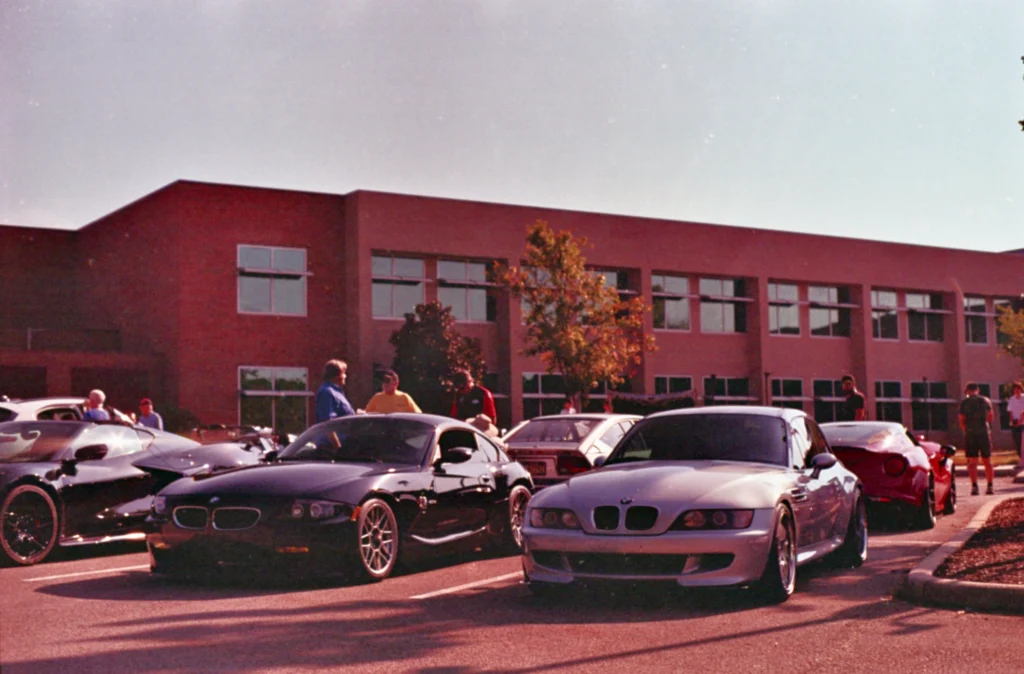 Black and silver BMW Z3 coupes parked in a lot with people and a red brick building in background during a car meet