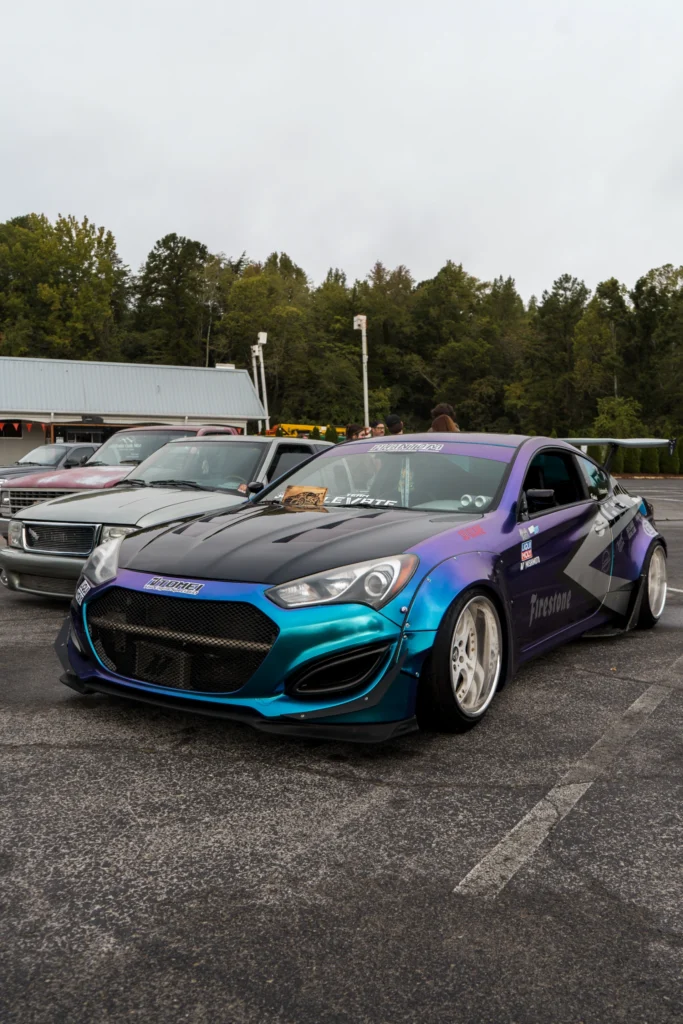 A purple and blue Hyundai Genesis Coupe with a black hood and rear wing sits beside pickups at an outdoor car meet.