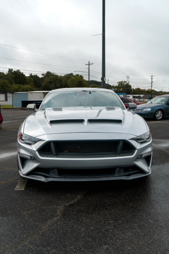 Silver Ford Mustang parked outdoors on wet asphalt with some buildings and other cars in background.