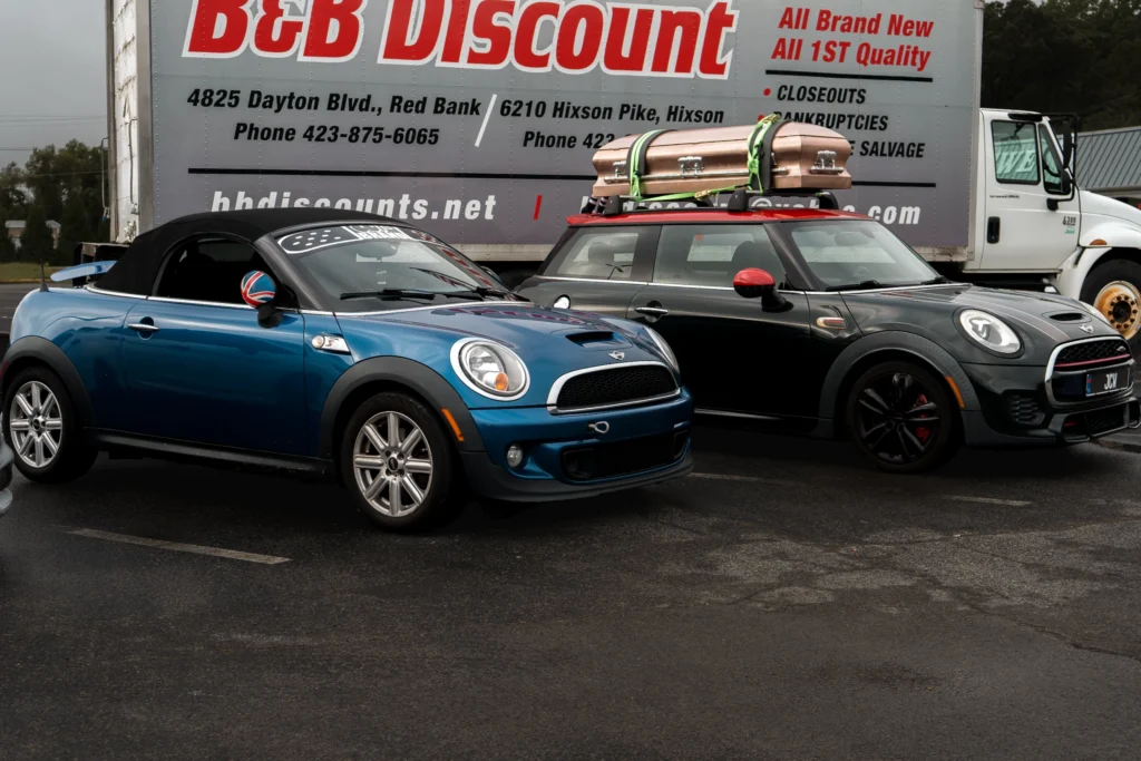 Blue Mini Cooper convertible and black Mini Cooper hardtop with red roof and roof rack carrying a casket, parked outdoors