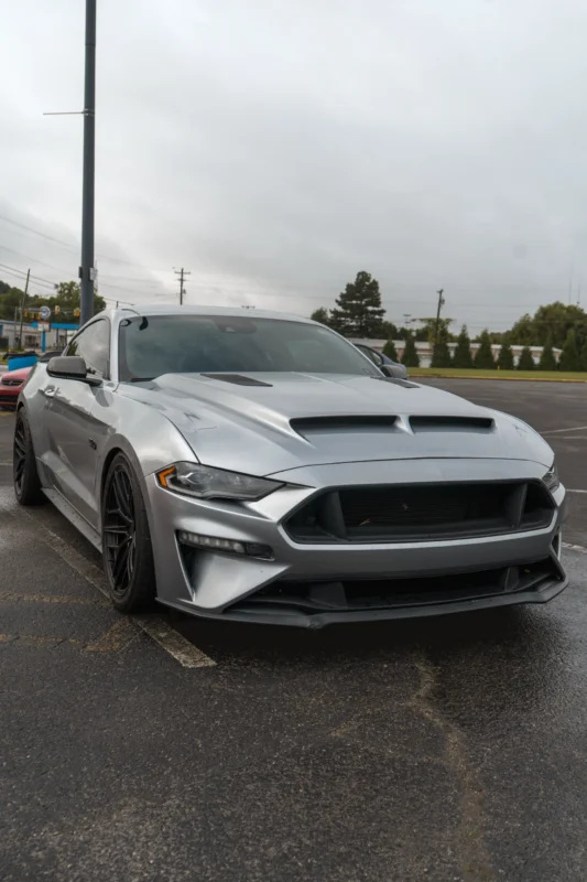 Silver Ford Mustang GT parked outdoors on an overcast day, facing front 3/4 with black wheels and visible hood vents.