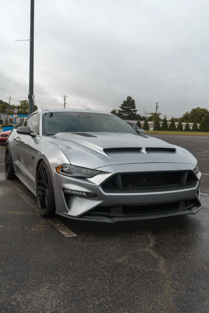 Silver Ford Mustang GT parked outdoors on an overcast day, facing front 3/4 with black wheels and visible hood vents.