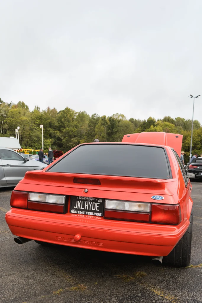 Red Ford Mustang parked at an outdoor car event with its hood open and other cars and people in the background.