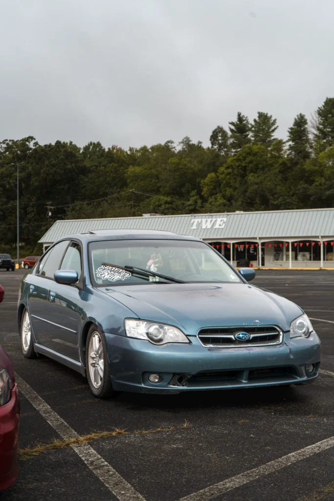 A blue Subaru Legacy sedan is parked outside a commercial building, viewed from the front three-quarter angle.