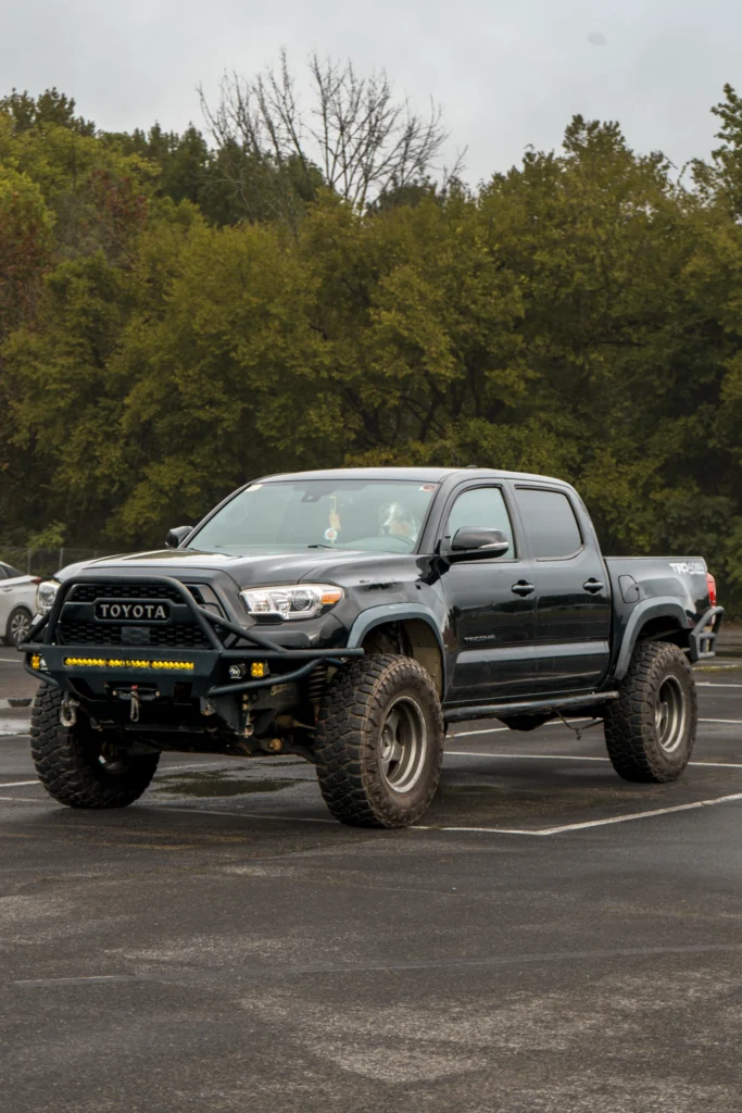 Black Toyota Tacoma pickup truck parked in a lot with trees in the background under cloudy sky